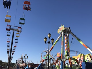 A busy amusement park with a bright blue sky in the background. A colorful chairlift full of people runs diagonally across the scene, with seats in red, yellow, green, and blue. Below, various rides and attractions, including a large swing ride and roller coaster tracks, are visible. Decorated lampposts are present in the foreground.
