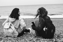 Two people sit on a pebbly beach, facing the sea, with the waves gently lapping in the background. They both have backpacks and appear to be engaged in conversation, exuding a sense of companionship and relaxation.