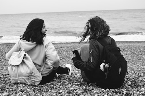 Two people sit on a pebbly beach, facing the sea, with the waves gently lapping in the background. They both have backpacks and appear to be engaged in conversation, exuding a sense of companionship and relaxation.