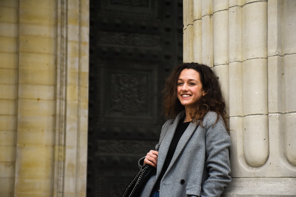 smiling woman holding black shoulder bag leaning on white wall