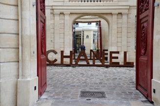 Large metal letters spelling 'Chanel' are positioned within an archway of a historic building, framed by open red doors on either side. The ground consists of cobblestones, and the building features classical architectural details and a window with a shop in the background.