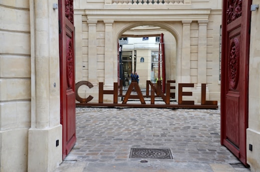 Large metal letters spelling 'Chanel' are positioned within an archway of a historic building, framed by open red doors on either side. The ground consists of cobblestones, and the building features classical architectural details and a window with a shop in the background.