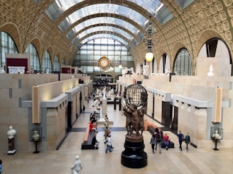 A grand interior of a museum with high, ornate ceilings and large arched windows, filled with sculptures and art pieces. People are walking and sitting around benches, absorbing the art surrounding them. A large bronze sculpture featuring human figures holding a globe is prominently displayed in the center.