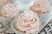 A close-up of delicate, pastel-colored cupcakes with creamy frosting, arranged on a cream tablecloth.