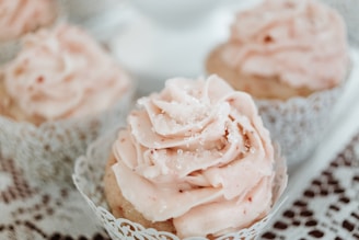 A close-up view of cupcakes with soft pink frosting, intricately piped on top and lightly sprinkled with sugar crystals. They are encased in delicate, lacy cupcake liners and set on a white surface.