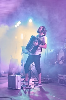 A musician playing an accordion on stage with a misty atmosphere and colorful lighting. The performer is wearing headphones and appears deeply engaged in the music. Surrounding equipment includes a drum set, lights, and cables.