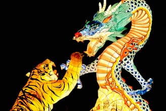 A vibrantly colored dragon and tiger lantern display against a black background. The dragon features intricate patterns with shades of blue, green, and orange, while the tiger is predominantly yellow with black stripes. Both figures are engaged in a dynamic pose, as though interacting or confronting each other.