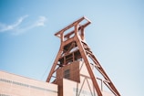 A large industrial structure with a distinctive red-brown metallic frame towers into the clear blue sky. The building is composed of geometric shapes, featuring a prominent tower with horizontal and diagonal beams. The structure is part of an industrial complex with a brick facade.