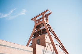 A large industrial structure with a distinctive red-brown metallic frame towers into the clear blue sky. The building is composed of geometric shapes, featuring a prominent tower with horizontal and diagonal beams. The structure is part of an industrial complex with a brick facade.