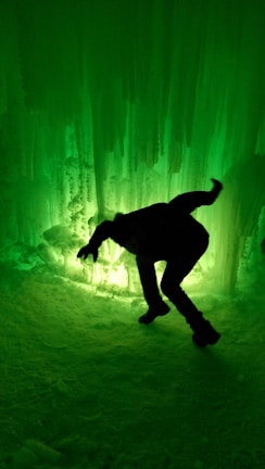 A person is in a bent position against an illuminated ice wall, creating a dramatic silhouette effect. The environment has an ethereal glow with varying shades of green, highlighting the icicles and snowy surface.