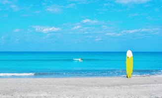 A serene beach scene with a calm, bright blue ocean under a partly cloudy sky. In the foreground, a vibrant yellow surfboard is standing upright on the sand. In the distance, a lone surfer is paddling along the water.
