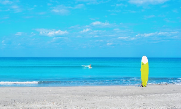 A serene beach scene with a calm, bright blue ocean under a partly cloudy sky. In the foreground, a vibrant yellow surfboard is standing upright on the sand. In the distance, a lone surfer is paddling along the water.