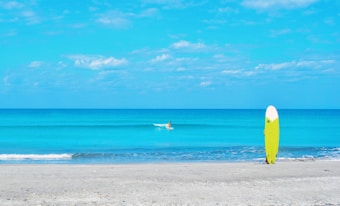 A serene beach scene with a calm, bright blue ocean under a partly cloudy sky. In the foreground, a vibrant yellow surfboard is standing upright on the sand. In the distance, a lone surfer is paddling along the water.