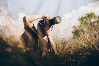 A person crouches in tall grass, holding a camera with a long lens to their eye, capturing a photograph. The scene appears to be set in a natural, outdoor environment with soft lighting and a serene backdrop of blurred trees and blue sky.
