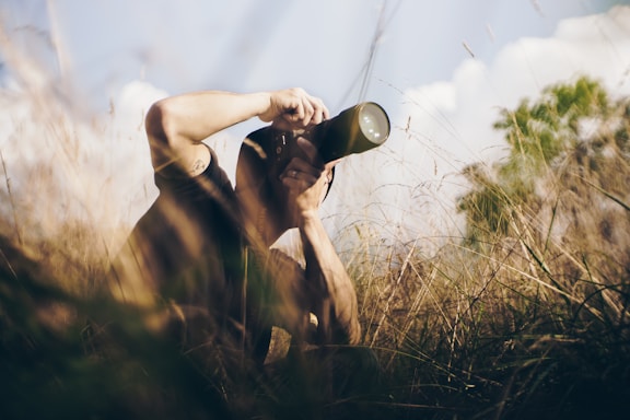 A person crouches in tall grass, holding a camera with a long lens to their eye, capturing a photograph. The scene appears to be set in a natural, outdoor environment with soft lighting and a serene backdrop of blurred trees and blue sky.