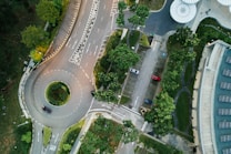 An aerial view of a roundabout intersected by multiple roads. Several cars are parked in the adjacent parking area. Greenery surrounds the roads, providing a lush and well-maintained look. A large building with a white roof is visible on the right side of the image, and the overall area appears organized with clear road markings.