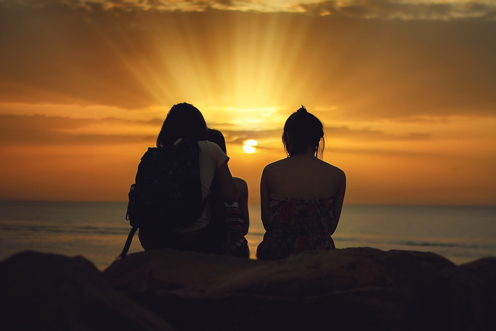 three person sitting on brown stone while watching the sunset