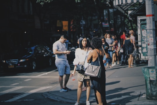 On a bustling city street, several people are captured in motion. Two women in the foreground engage in conversation, one carrying a purse. In the background, more passersby walk along the sidewalk, partially obscured by shadows and urban elements like cars, a garbage bin, and shop awnings.