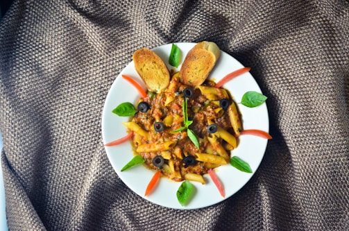 A plate of penne pasta with a rich meat sauce, garnished with black olives, green basil leaves, and slices of red tomato. Two toasted bread slices are placed at the top of the plate. The presentation is on a textured brown fabric.