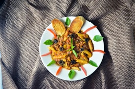 A plate of penne pasta with a rich meat sauce, garnished with black olives, green basil leaves, and slices of red tomato. Two toasted bread slices are placed at the top of the plate. The presentation is on a textured brown fabric.