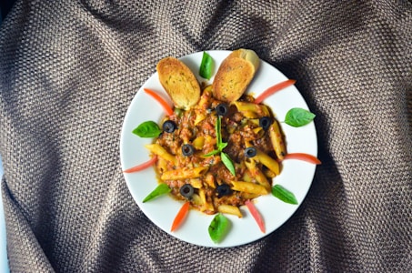 A plate of penne pasta with a rich meat sauce, garnished with black olives, green basil leaves, and slices of red tomato. Two toasted bread slices are placed at the top of the plate. The presentation is on a textured brown fabric.