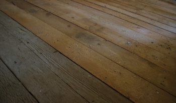 A close-up view of an aged, wooden floor with visible planks. The surface appears worn, with some dust and debris scattered across the boards.
