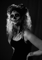 A person with dramatic makeup resembling a skull, featuring intricate designs on the face. The hair is styled with volume and curls, and the image is in black and white, accentuating the contrast of the makeup. The person is wearing a sleeveless dark top and has visible tattoos on the arm.