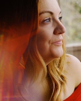 A woman with long, light brown hair gazes thoughtfully out of a window. She is wearing an off-the-shoulder red top. The image has a soft and warm appearance, with a blurred orange-red reflection or flare effect on the left side.