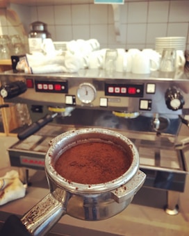 A close-up view of a portafilter filled with ground coffee held in front of a large espresso machine. In the background, several stacked white cups and other coffee-making equipment are placed on a tiled countertop.