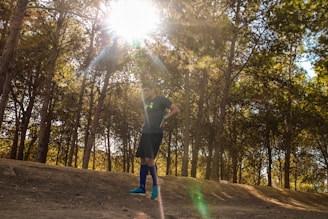 A person wearing a sports outfit stands under tall trees with sunlight streaming through the branches, casting dappled shadows on the dirt path. The individual appears to be preparing for a run or a workout.