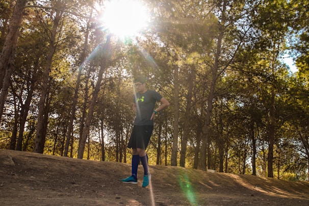 A person wearing a sports outfit stands under tall trees with sunlight streaming through the branches, casting dappled shadows on the dirt path. The individual appears to be preparing for a run or a workout.