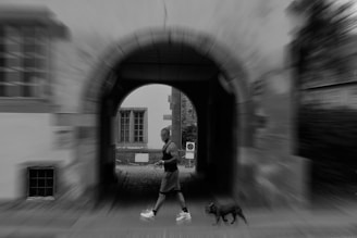 A person is jogging through an arched passageway with a small dog on a leash. The surrounding area is blurred, suggesting motion. The scene appears to be in a historic or old town setting, with cobblestone and traditional architecture.