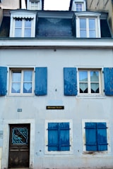 A quaint two-story building with a slate roof and dormer windows at the top. Blue wooden shutters frame the windows, adding an old-world charm. The building's facade is painted in pale hues, giving it a weathered yet inviting appearance. A small plaque labeled 'Poissonnerie' suggests this might be a fish shop.