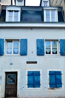 A quaint two-story building with a slate roof and dormer windows at the top. Blue wooden shutters frame the windows, adding an old-world charm. The building's facade is painted in pale hues, giving it a weathered yet inviting appearance. A small plaque labeled 'Poissonnerie' suggests this might be a fish shop.