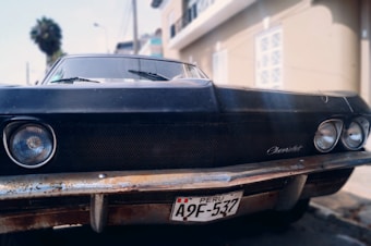 A close-up view of the front of an old, black Chevrolet car with some rust visible on the bumper. The car is parked on a street with a beige building in the background. The license plate is from Peru and reads A9F-537.