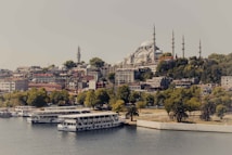 A picturesque view of a cityscape with a large mosque featuring multiple minarets atop a hill, surrounded by lush greenery and residential buildings. The foreground showcases a tranquil body of water with several docked boats.
