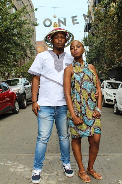 A man and a woman stand confidently on a city street with parked cars on either side. The man wears a white shirt with traditional patterns and blue jeans, accessorized with a colorful hat. The woman is dressed in a vibrant patterned sleeveless dress and sandals. Behind them, trees line the street, and there is a visible overhead sign with letters.
