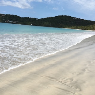 A serene beach scene features gentle waves washing onto a sandy shore. The turquoise water meets the soft sand, creating a peaceful and inviting atmosphere. In the background, lush green hills and distant white sailboats blend with the clear blue sky, dotted with a few white clouds.