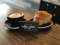 A cup of frothy coffee in a black cup and saucer is placed on a wooden table next to a golden-brown croissant. Between them stands a black sign card with white text that reads 'Irving Park Coffee Joint, Coffee Tea Sandwiches.' A chair with a black backrest is partially visible in the background.