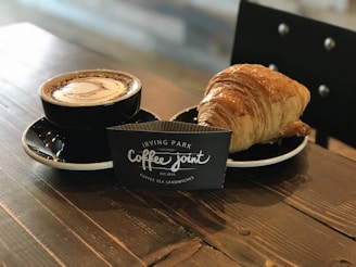 A cup of frothy coffee in a black cup and saucer is placed on a wooden table next to a golden-brown croissant. Between them stands a black sign card with white text that reads 'Irving Park Coffee Joint, Coffee Tea Sandwiches.' A chair with a black backrest is partially visible in the background.