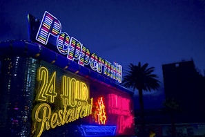 Bright neon signs are illuminated against a nighttime backdrop, displaying 'Peppermill' and '24 Hour Restaurant' in vibrant colors. There is a palm tree silhouetted against the deep blue sky.