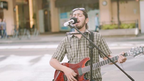 A musician performs on the street, passionately singing into a microphone. The person is playing an electric guitar and has a harmonica attached to a holder around their neck. They are dressed in a plaid shirt and sunglasses resting on their head, while a blurred urban background suggests a public space.