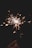 A bright sparkler is held against a dark background, emitting sparks in all directions. The sparks create a dynamic and shimmering effect, illuminating the surroundings with a warm glow.