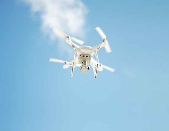 A white quadcopter drone is flying against a clear blue sky with some subtle clouds. The drone's propellers are in motion, and its camera is clearly visible on the underside.