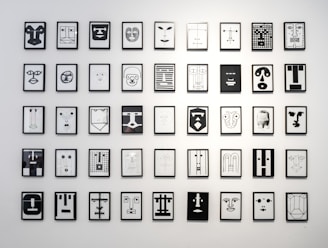 A collection of framed abstract line art drawings is hung in a grid pattern on a white wall. Each drawing features geometric designs resembling stylized faces, using minimalistic lines and shapes in black and white.