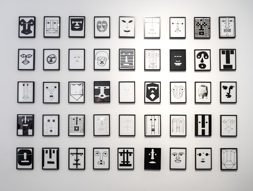 A collection of framed abstract line art drawings is hung in a grid pattern on a white wall. Each drawing features geometric designs resembling stylized faces, using minimalistic lines and shapes in black and white.