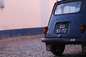 The image captures the rear side of a vintage blue car with a black license plate that reads 'GQ 51-72.' The car is parked on a cobblestone street next to a tall wall that appears to be painted in white with a blue border at the bottom. The car's window has several stickers, including one with a camel and another with a peace sign. The car is slightly rusted indicating its age.
