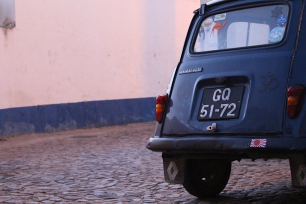 The image captures the rear side of a vintage blue car with a black license plate that reads 'GQ 51-72.' The car is parked on a cobblestone street next to a tall wall that appears to be painted in white with a blue border at the bottom. The car's window has several stickers, including one with a camel and another with a peace sign. The car is slightly rusted indicating its age.