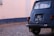 The image captures the rear side of a vintage blue car with a black license plate that reads 'GQ 51-72.' The car is parked on a cobblestone street next to a tall wall that appears to be painted in white with a blue border at the bottom. The car's window has several stickers, including one with a camel and another with a peace sign. The car is slightly rusted indicating its age.