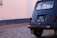 The image captures the rear side of a vintage blue car with a black license plate that reads 'GQ 51-72.' The car is parked on a cobblestone street next to a tall wall that appears to be painted in white with a blue border at the bottom. The car's window has several stickers, including one with a camel and another with a peace sign. The car is slightly rusted indicating its age.